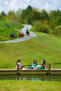 Group of three girls on the dock in the foreground, with three horseback riders on a path in the distance, waving