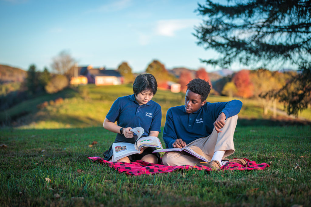 Two students sitting outside on picnic blanket, looking at textbook
