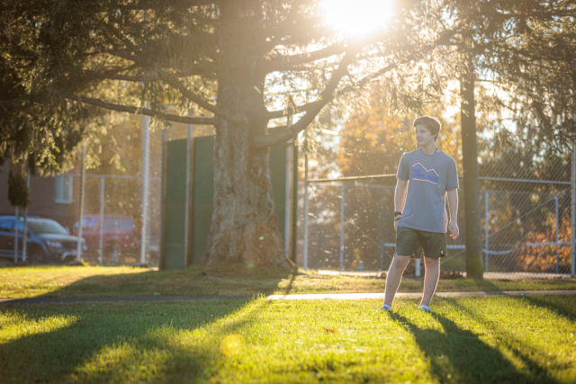 Student standing on grass in front of sports court with sunlight behind.